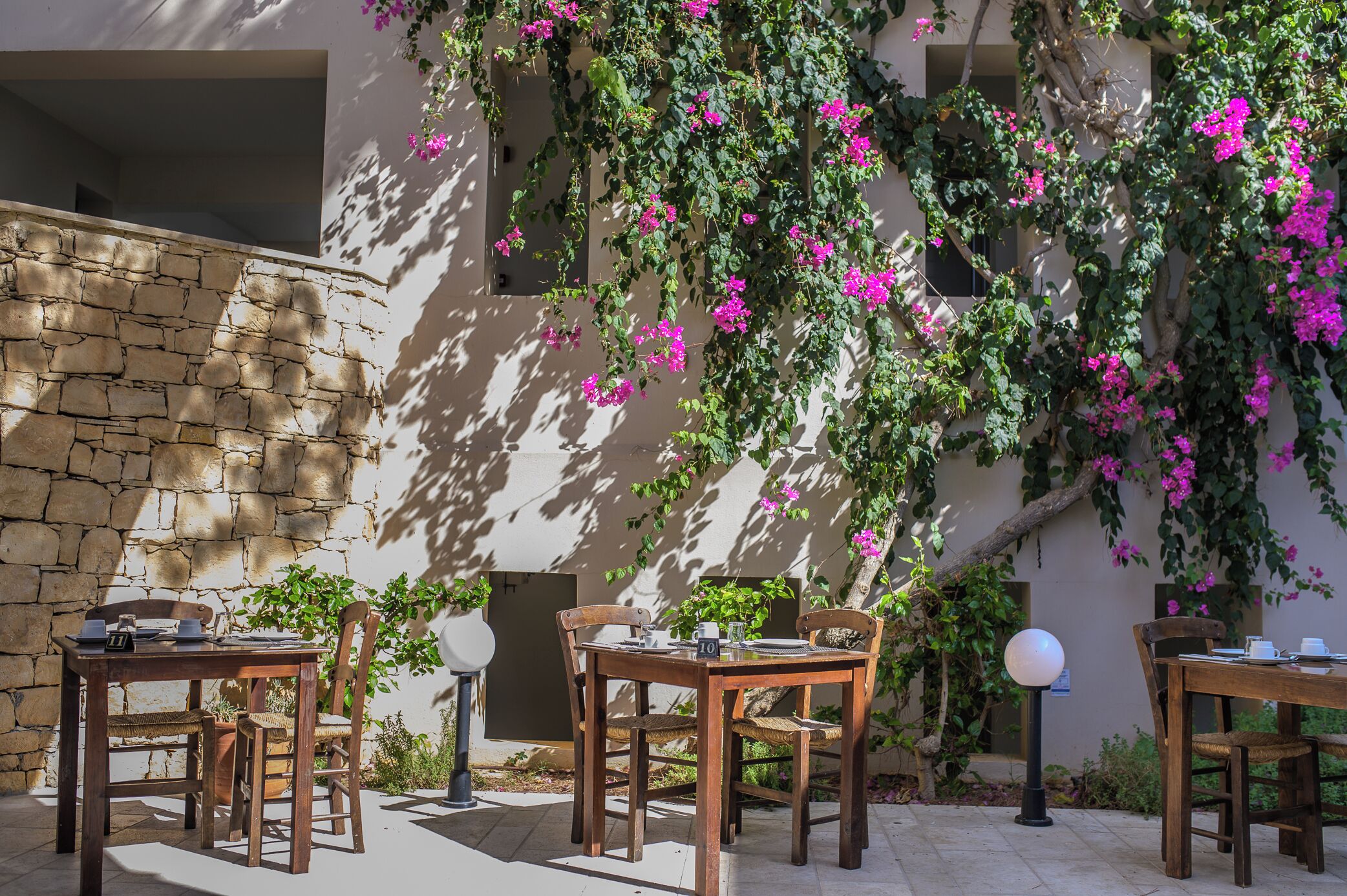 An outdoor dining area with stone walls, wooden tables, and chairs, surrounded by lush greenery and flowering vines.