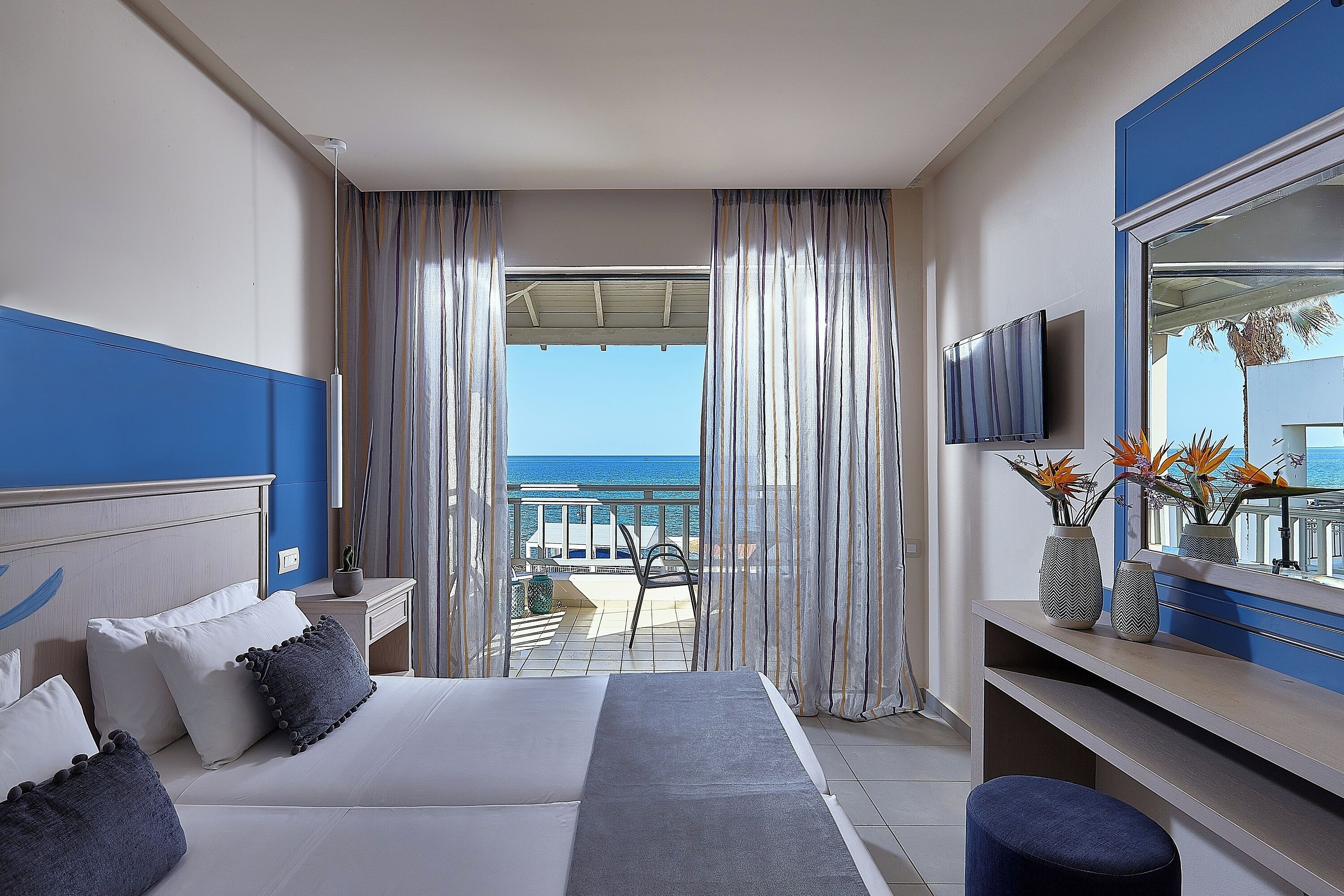 A hotel room with a bed, a desk, a TV, and a balcony with a view of the sea.