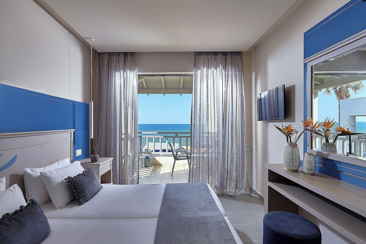 A hotel room with a bed, a desk, a TV, and a balcony with a view of the sea.