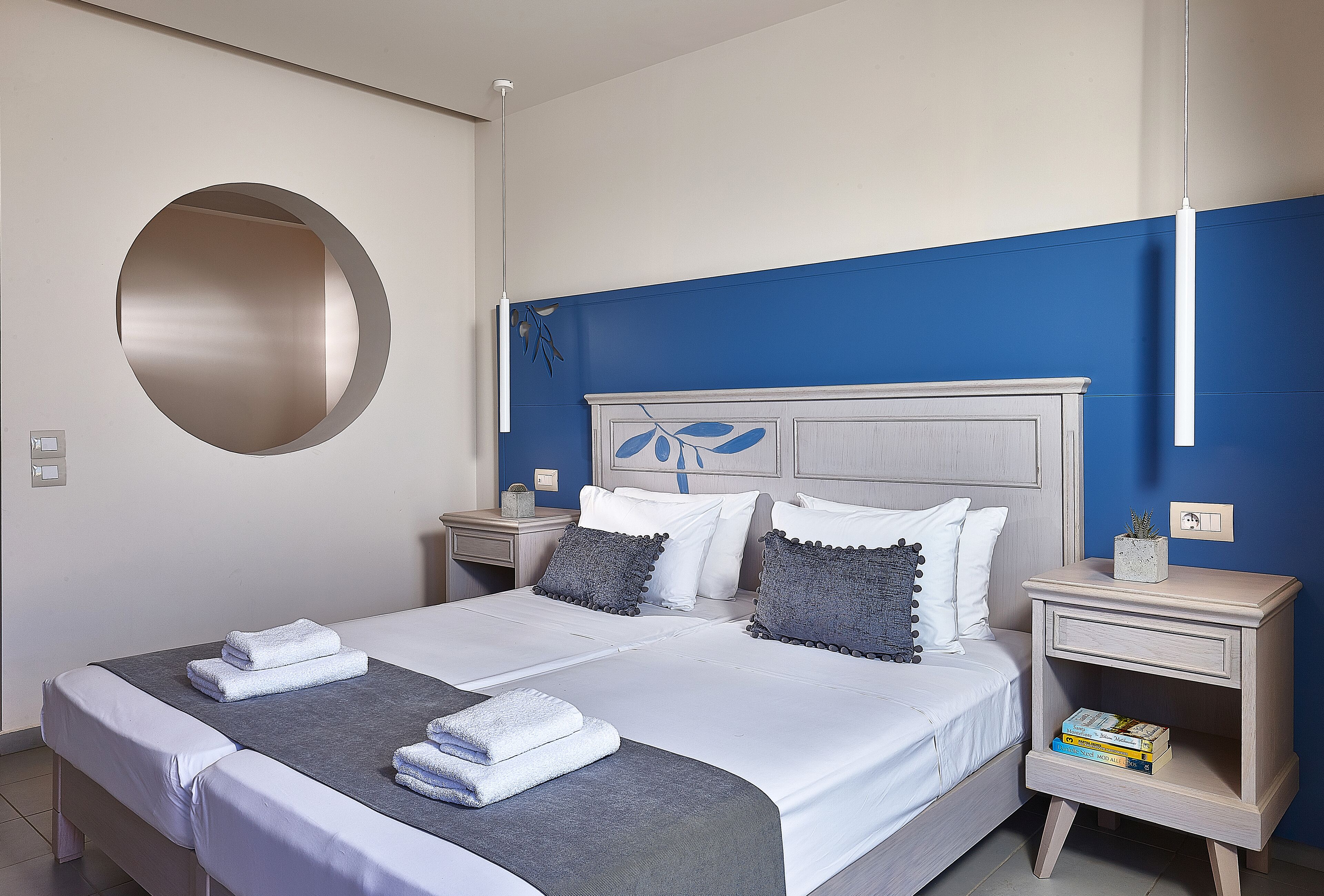 A neatly made bed with white linens and two folded towels, a bedside table with a book, and a wall-mounted light fixture.