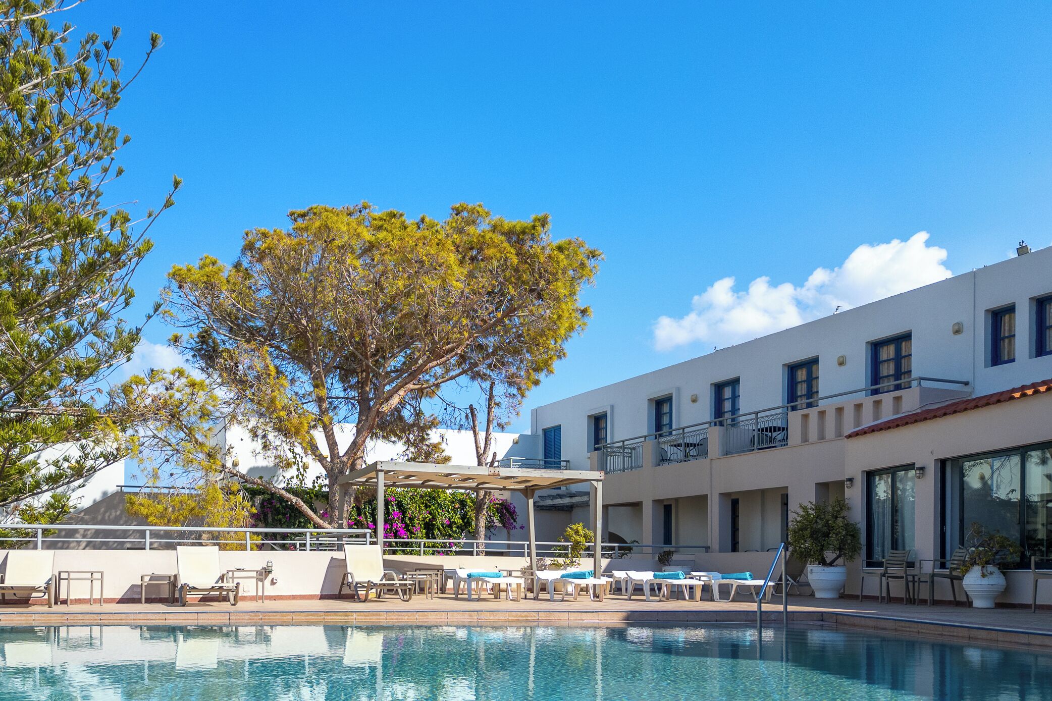 Un espace piscine agrémenté de chaises longues et d’une pergola, entouré d’arbres et bordé d’un bâtiment doté de balcons.