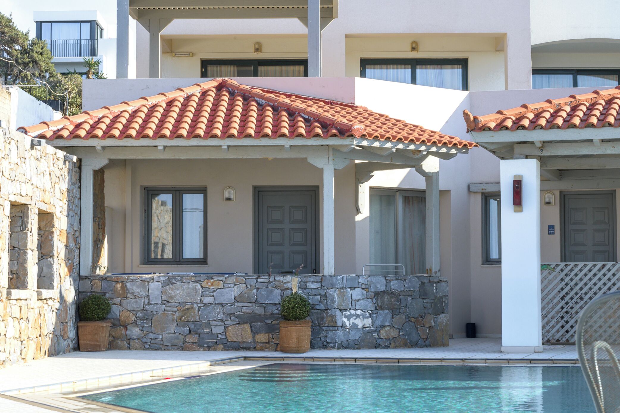 A poolside area with a stone wall, a covered patio, and a building with a tiled roof.