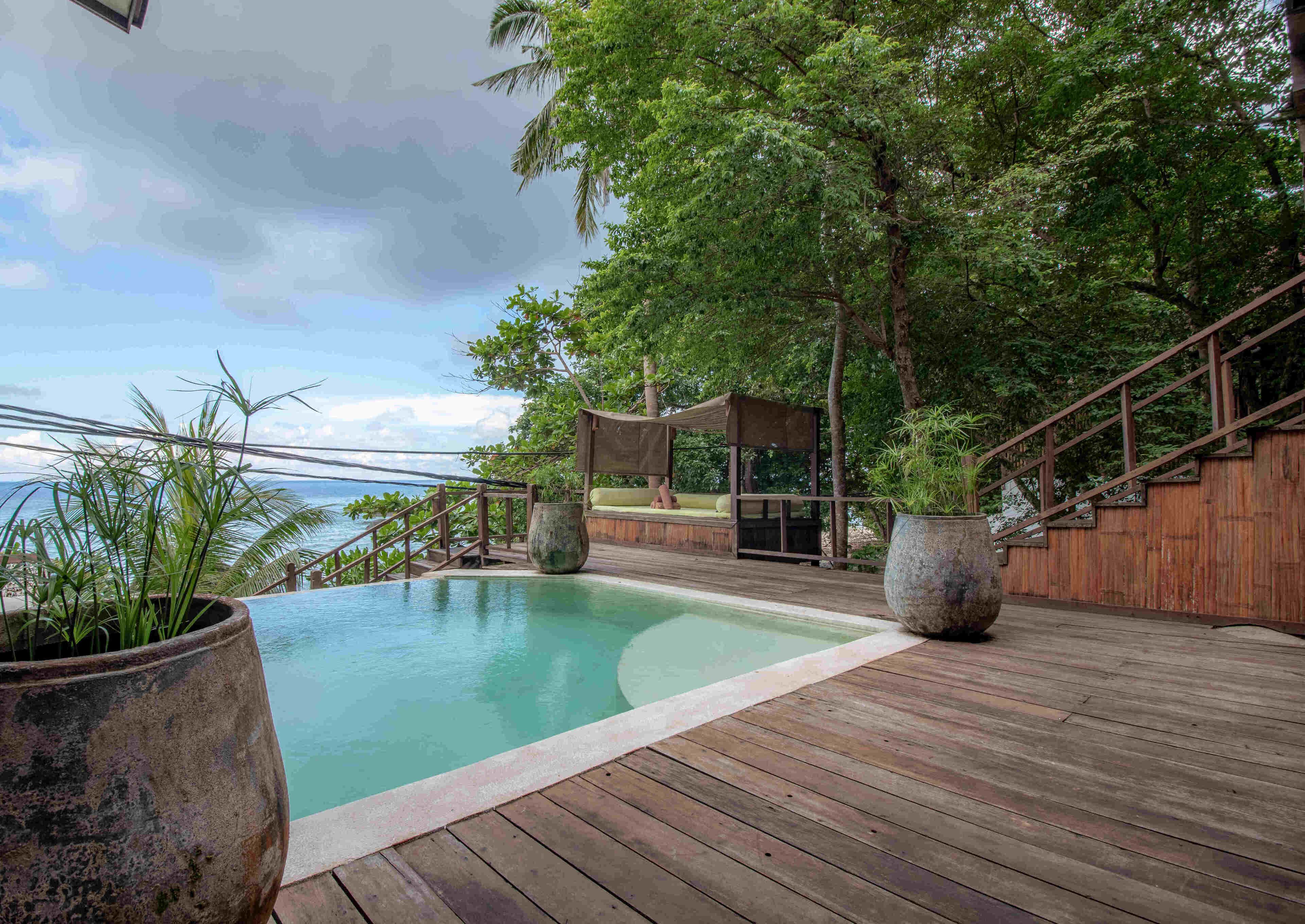 A wooden deck with a pool, surrounded by lush greenery and a view of the ocean.