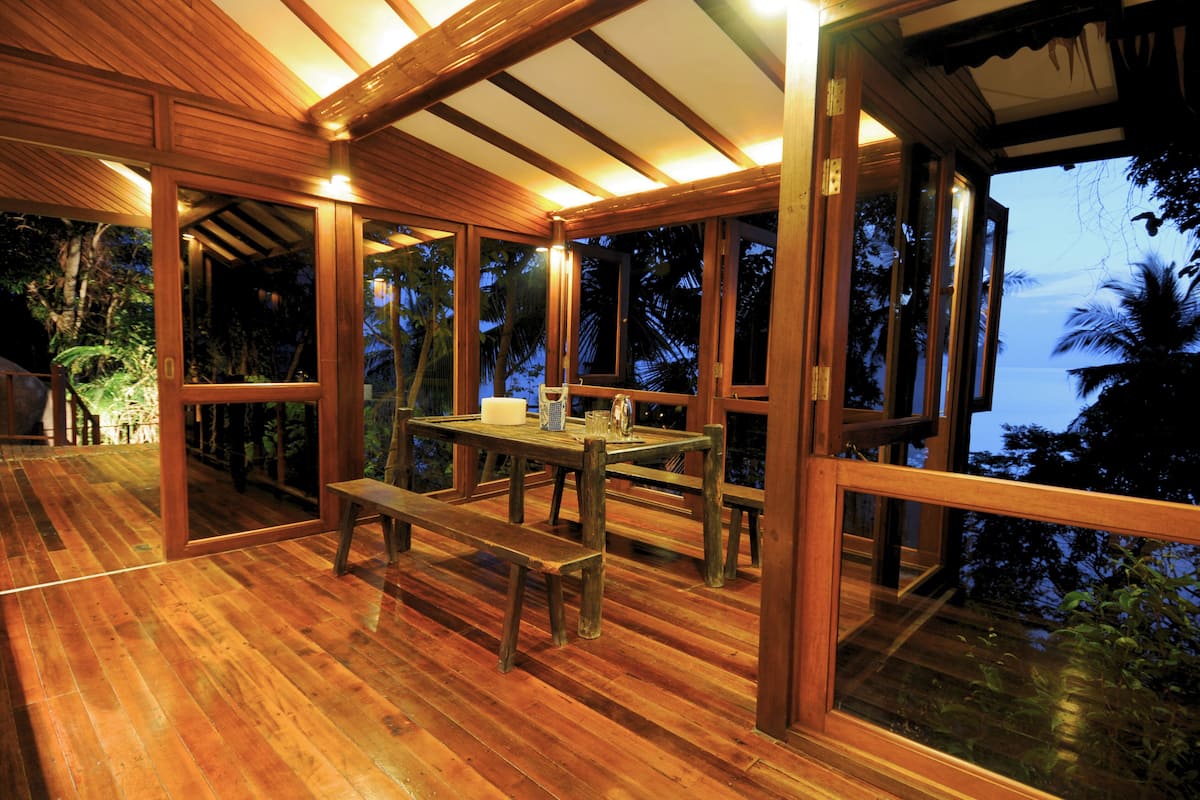 A wooden deck with a table and benches, large windows, and a view of the ocean.