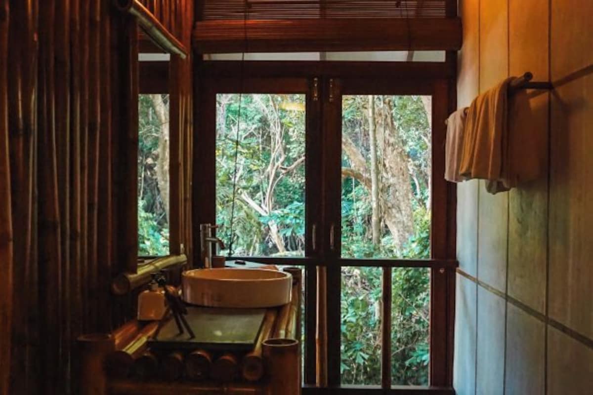 A rustic bathroom with a wooden sink, a mirror, and a view of dense foliage through large windows.