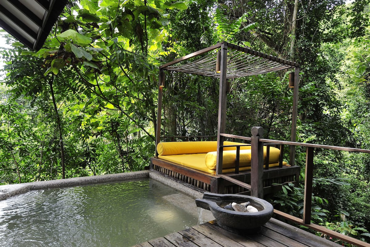 A wooden deck with a hot tub and a yellow cushioned seating area under a wooden pergola surrounded by lush greenery.