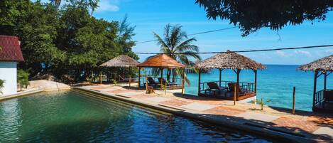 A poolside area with lounge chairs and thatched umbrellas, overlooking a clear blue sea.