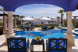 A pool area with blue chairs and umbrellas, a white building with a dome in the background, and palm trees.