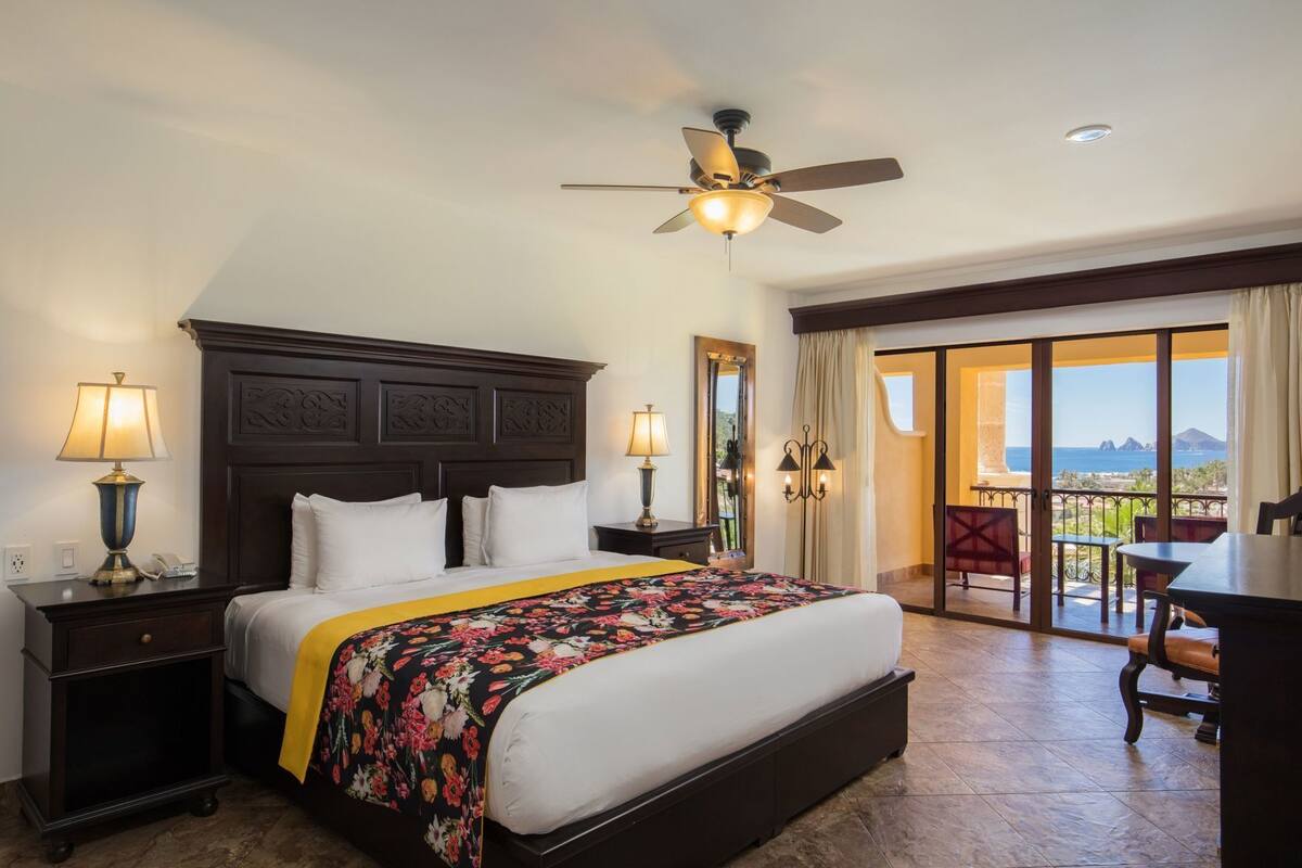 Quarto de hotel com uma cama grande, ventoinha no teto, varanda com vista e escrivaninha com cadeira.