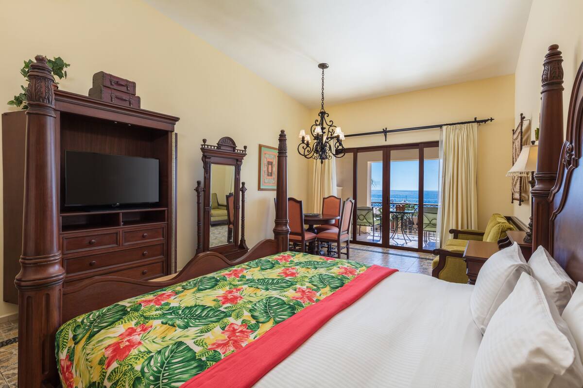 Quarto de hotel com uma cama grande, televisão, cômoda de madeira e varanda com vista para o oceano.