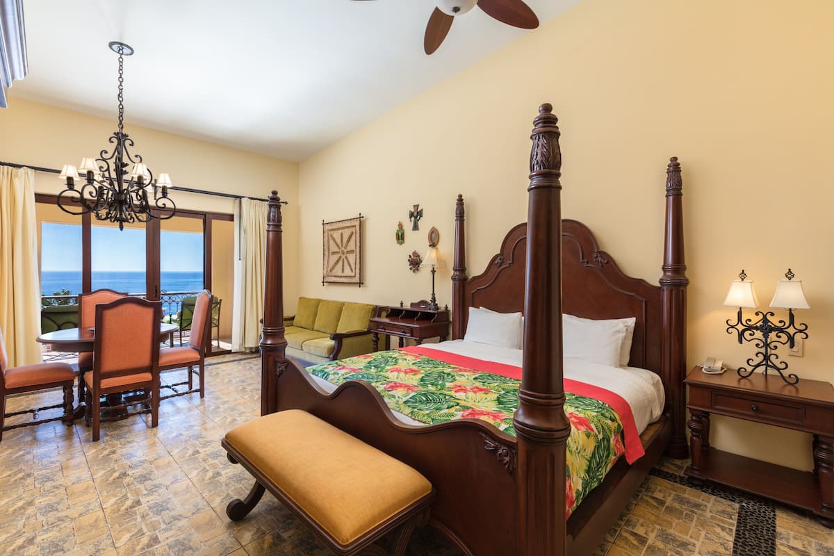 Uma cama de dossel com colcha florida, um banco, um candelabro e vista para o oceano.