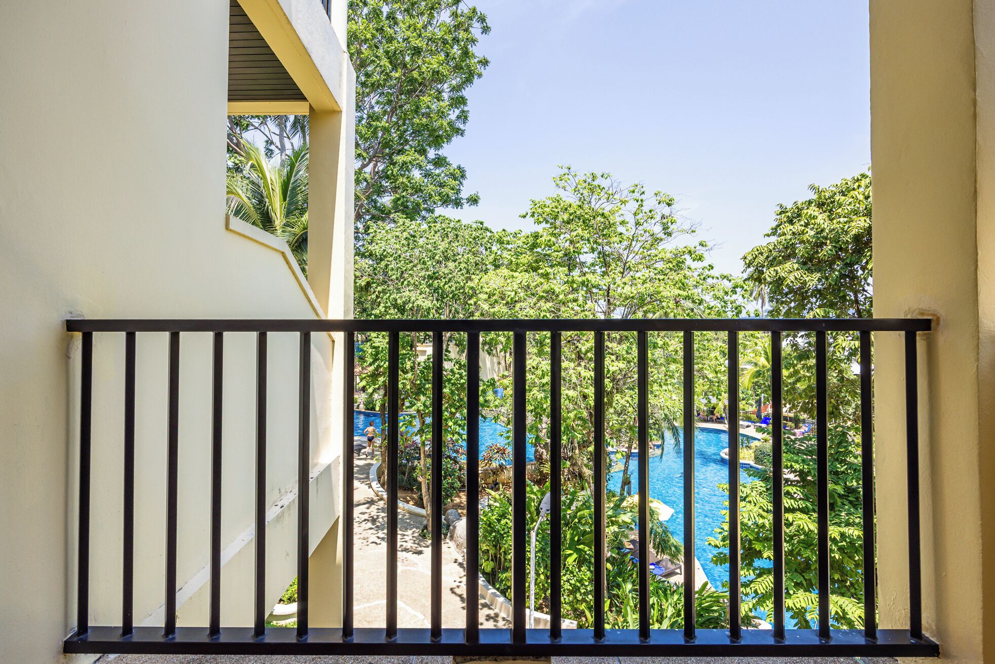 A balcony with a black metal railing overlooking a swimming pool surrounded by lush greenery.