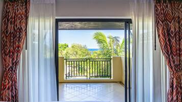 A room with a bed, a balcony with a view of the ocean, and patterned curtains.