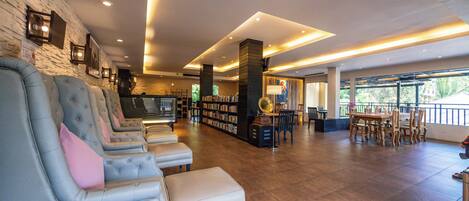 A hotel lobby with a seating area, a bookshelf, a dining table, and a large window.