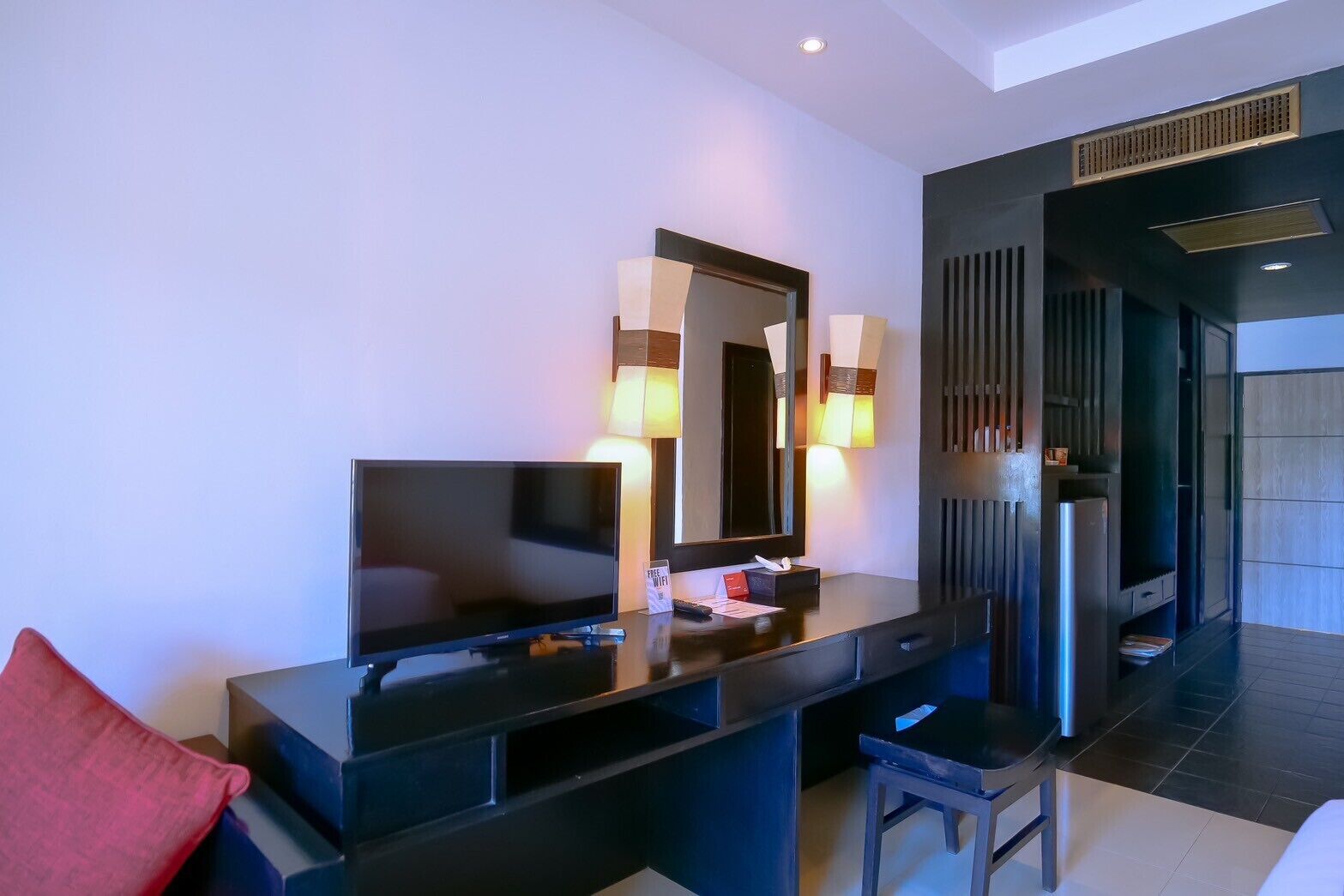 Habitación de hotel moderna con un televisor de pantalla plana grande, un tocador de madera oscura con espejo y un cojín rojo.