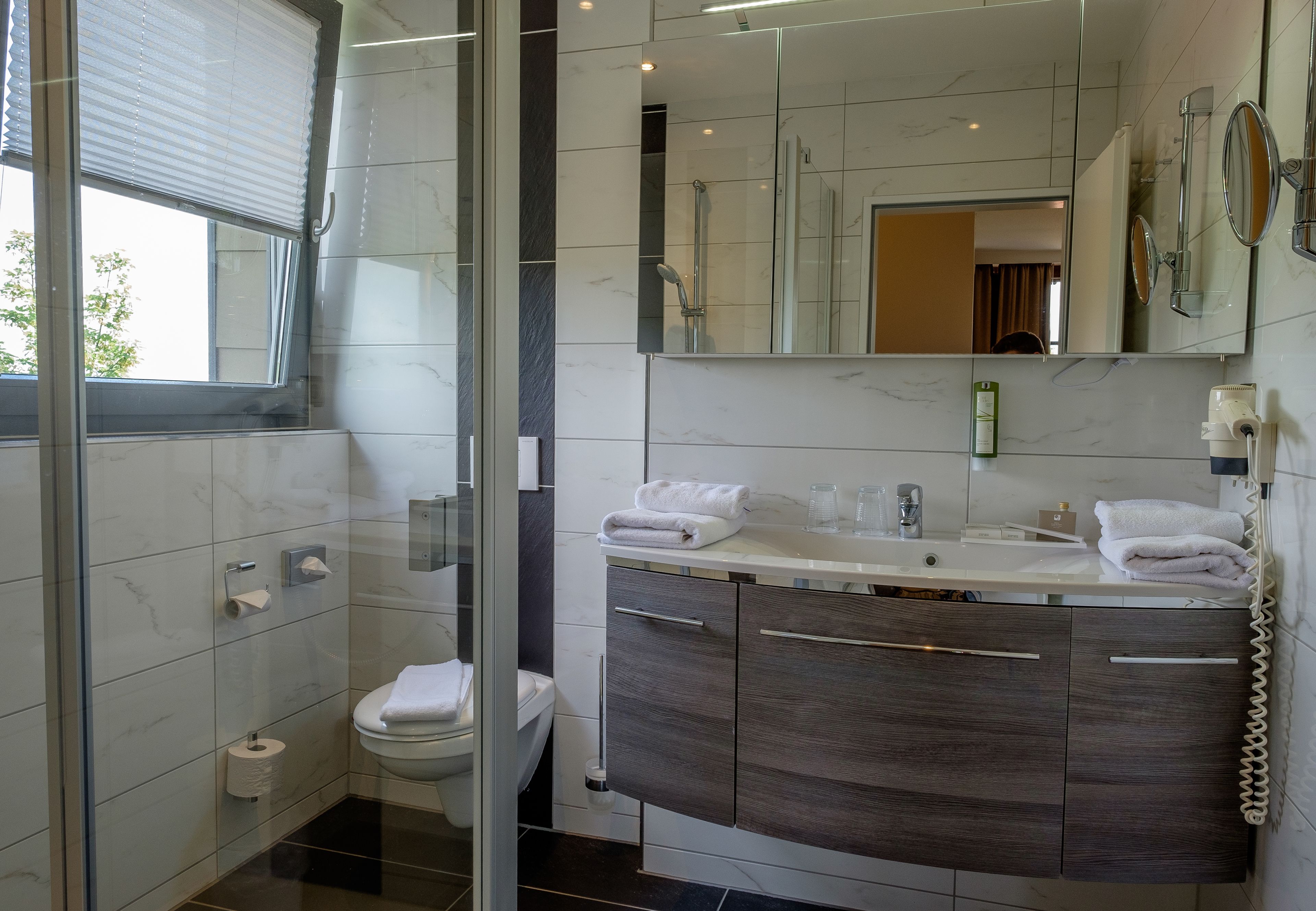 Un baño moderno con cabina de ducha de vidrio, inodoro, tocador con espejo y ventana con persianas.