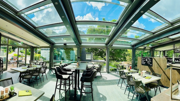 Ein Restaurant mit Glasdach, Außenplätzen und Blick auf Himmel und Bäume.