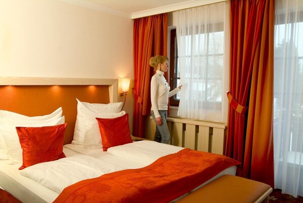 Habitación de hotel con cama, almohadas rojas y blancas, ventana con cortinas rojas y vista a árboles.