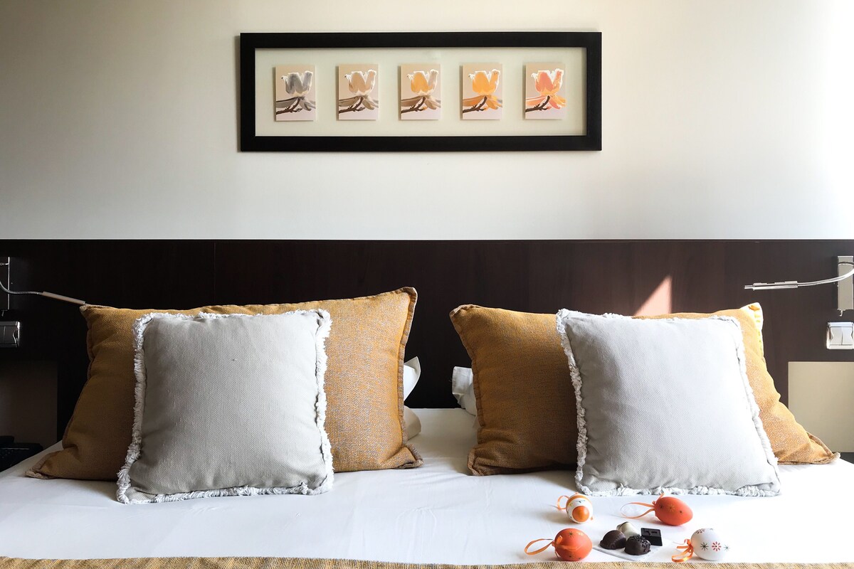 A neatly made bed with four pillows, a framed artwork above, and a bedside table with a lamp.