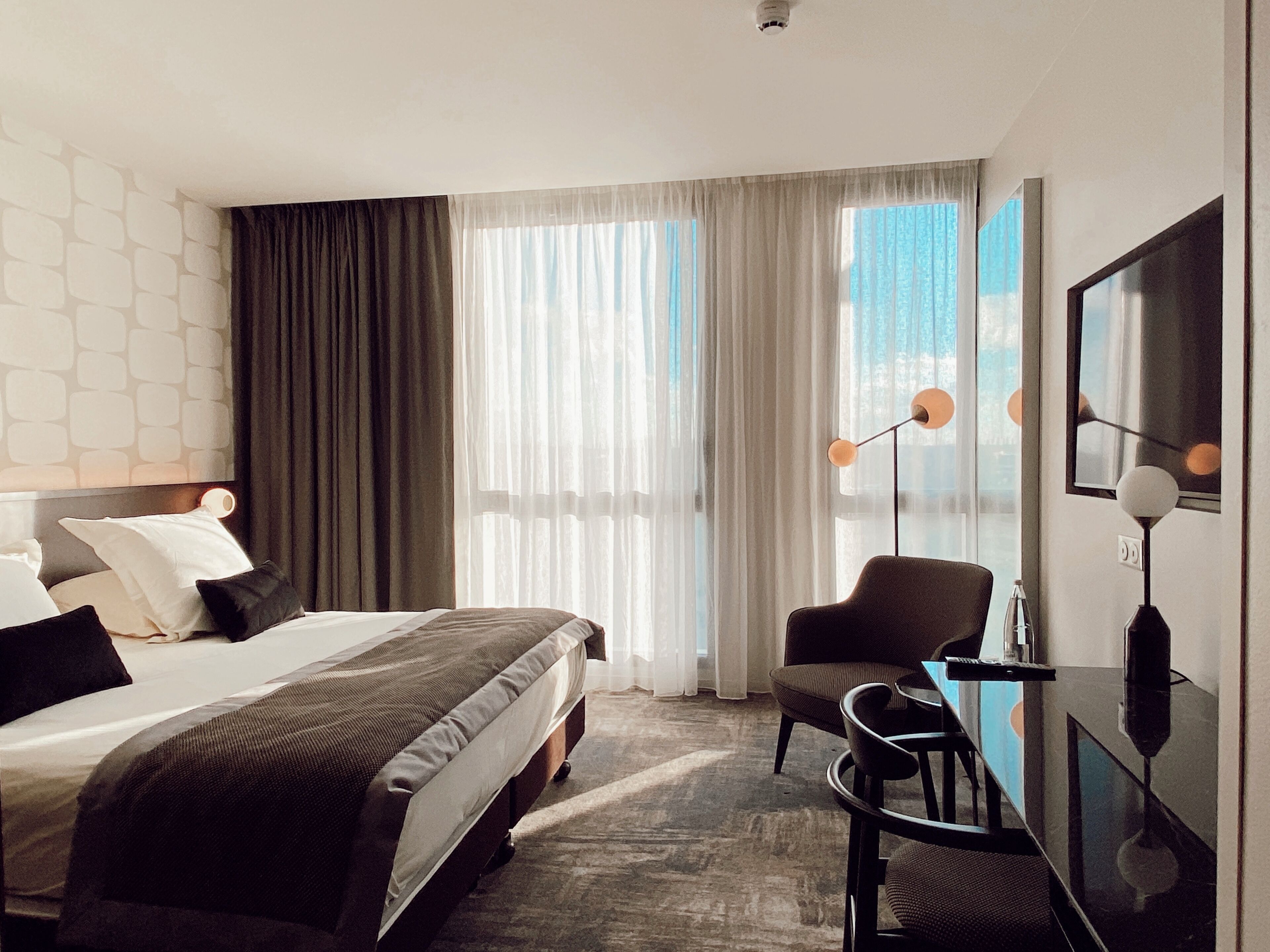 Une chambre d’hôtel moderne avec un grand lit, un bureau et une chaise. Il y a une fenêtre avec des rideaux légers et une vue sur l’extérieur.