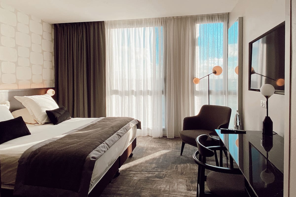 Une chambre d’hôtel moderne avec un grand lit, un bureau et une chaise. Il y a une fenêtre avec des rideaux légers et une vue sur l’extérieur.