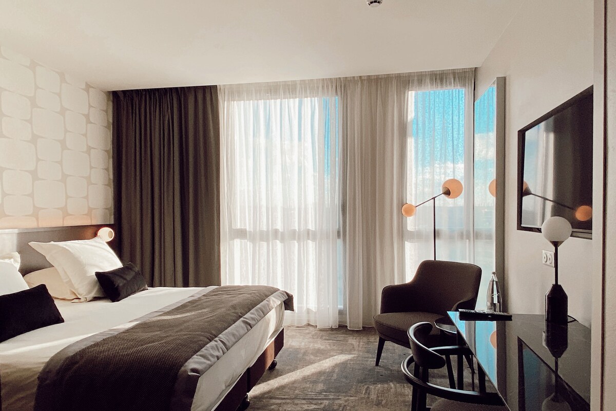 Ein modernes Hotelzimmer mit einem großen Bett, einem Schreibtisch und einem Stuhl. Ein Fenster mit durchscheinenden Vorhängen bietet einen Blick ins Freie.