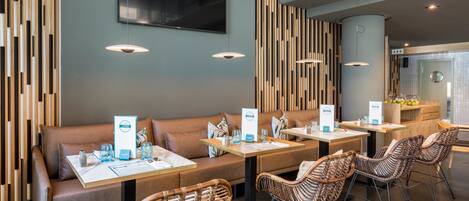 A modern restaurant with wooden panel walls, a flat-screen TV, and wicker chairs. Tables have menus and glasses.