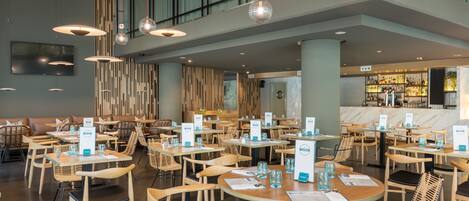 Ein modernes Restaurant mit runden Tischen und Holzstühlen, ein Barbereich im Hintergrund und Pendelleuchten.