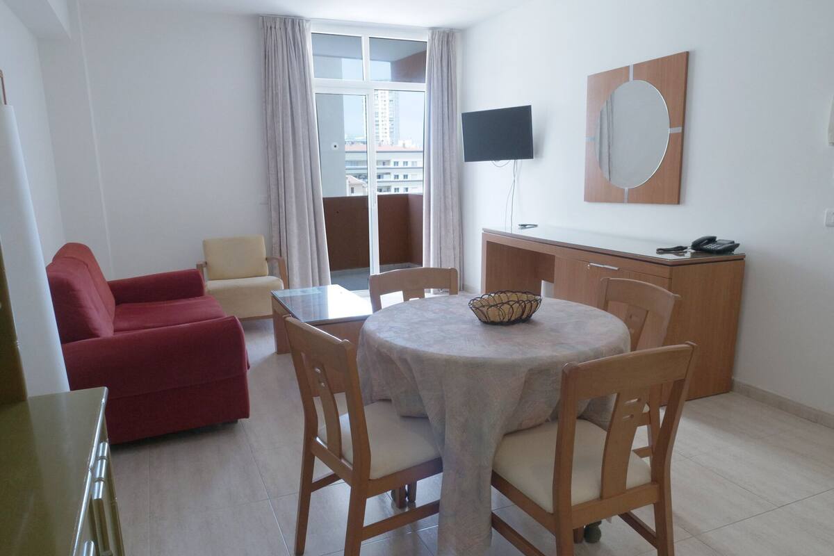 A modern living room with a red sofa, a beige sofa, a round dining table with a white tablecloth, wooden chairs, a TV mounted on the wall, and a wooden cabinet with a phone on top.