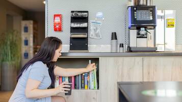A woman in a light blue dress reaching for books from a wooden shelf in a modern, well-lit room with a coffee machine and a red fire extinguisher on the wall.