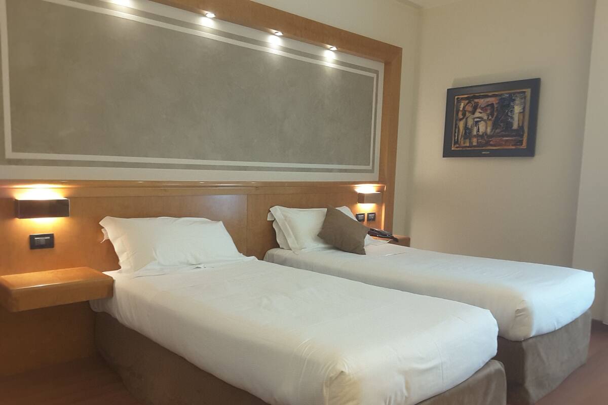 A hotel room with two single beds, wooden headboards, bedside lamps, a painting on the wall, and a large window.
