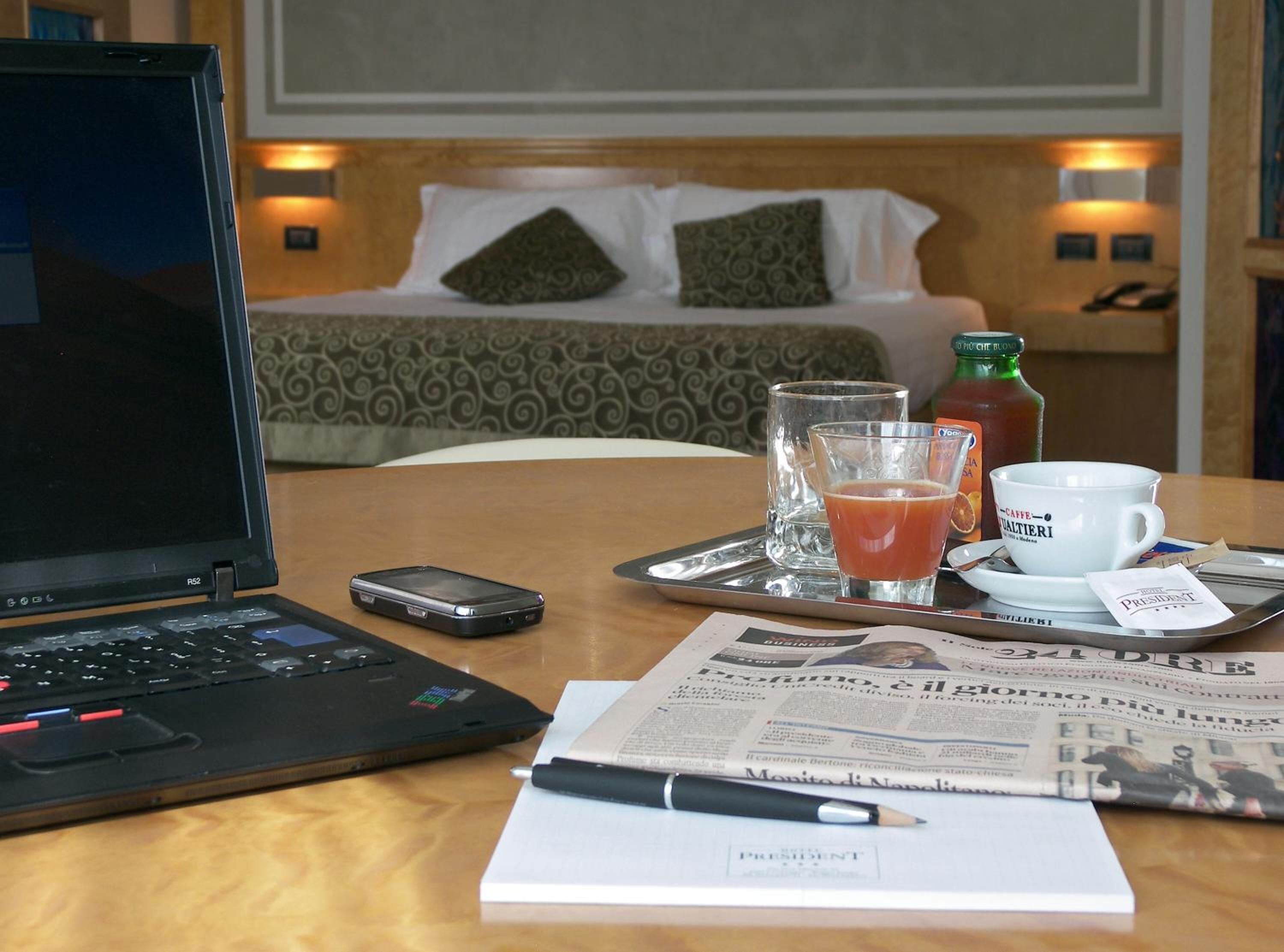 Ein Hotelzimmer mit einem Bett, einem Laptop, einem Smartphone, einem Tablett mit einem Glas Saft, einer Tasse Kaffee und einer Zeitung.