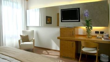 Et hotelværelse med en seng, et skrivebord med stol, et fjernsyn, en plante og et billede på væggen.