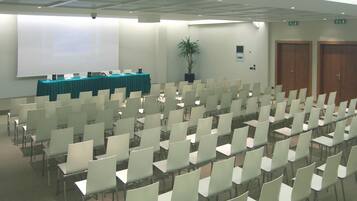 Een congreszaal met rijen stoelen, een scherm voor presentaties, een tafel met een groene tafelkleed en een potplant.