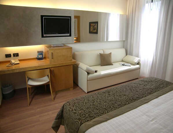 Habitación de hotel con escritorio, sofá, cama, televisor y ventana con cortinas.