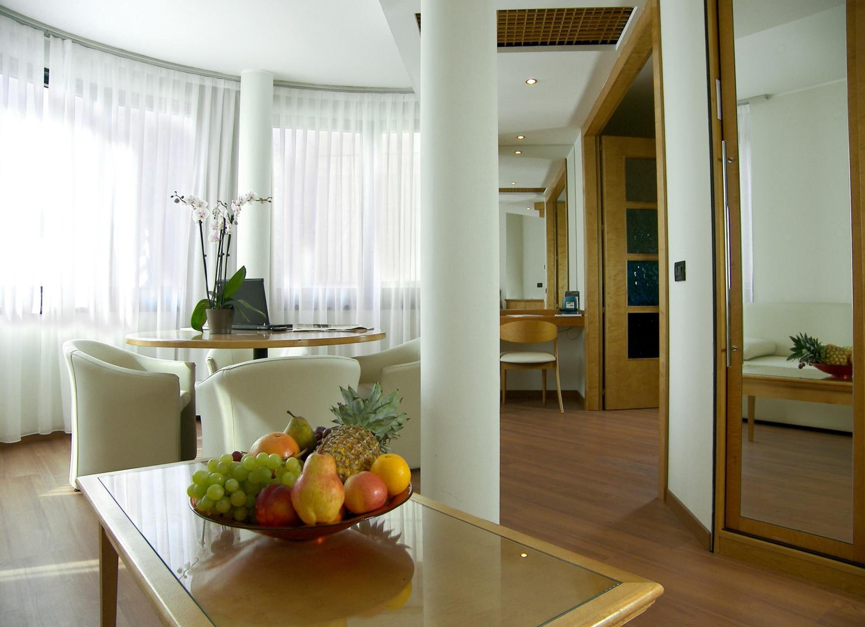 A modern hotel room with a dining area featuring a glass table with a fruit bowl, white chairs, and a wooden desk.