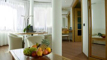 A modern hotel room with a dining area featuring a glass table with a fruit bowl, white chairs, and a wooden desk.
