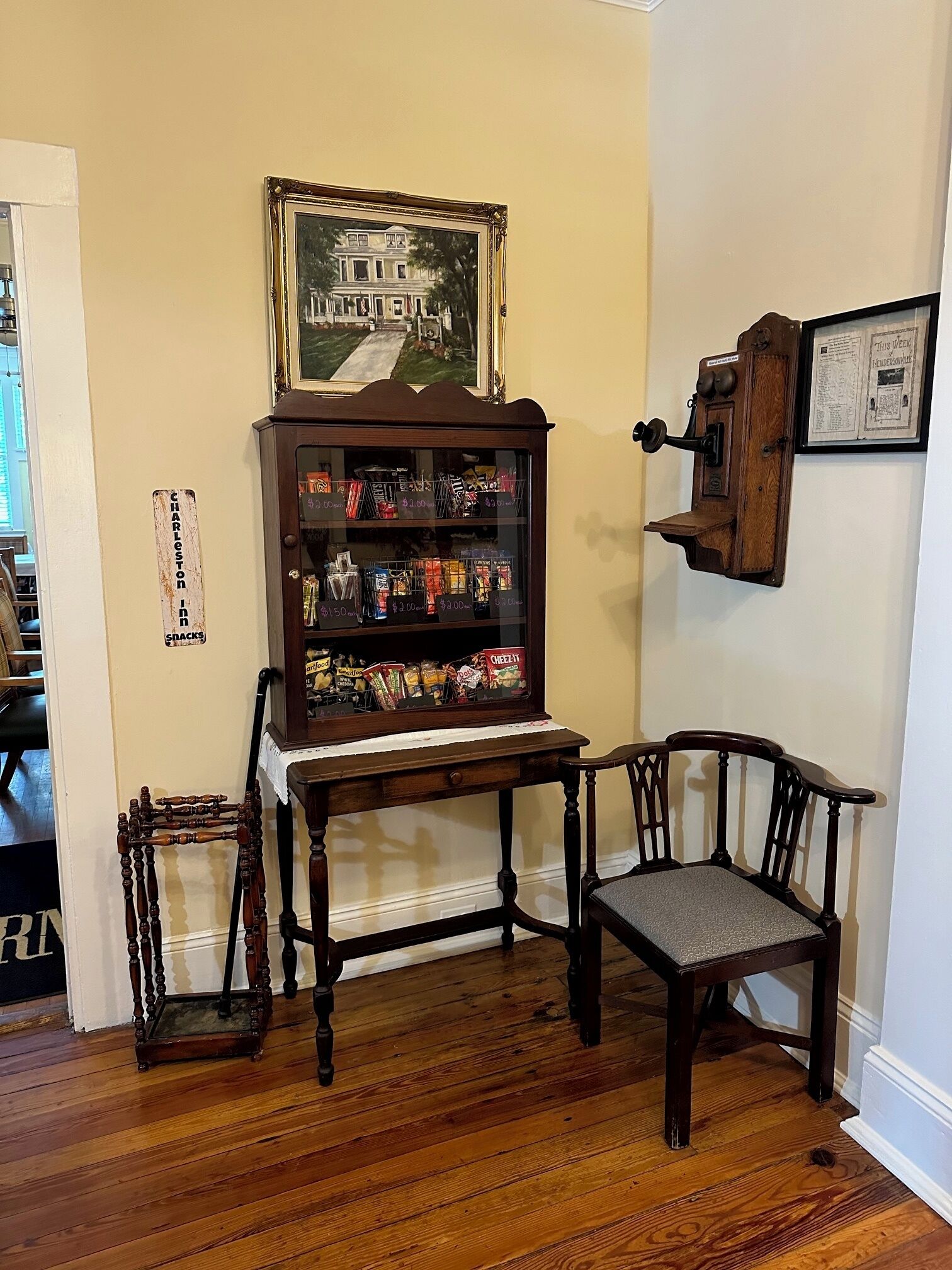 A room with a wooden cabinet displaying snacks, a framed picture above it, and a wooden chair.