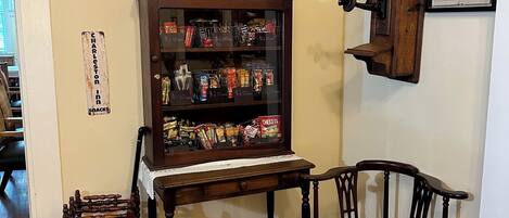A room with a wooden cabinet displaying snacks, a framed picture above it, and a wooden chair.