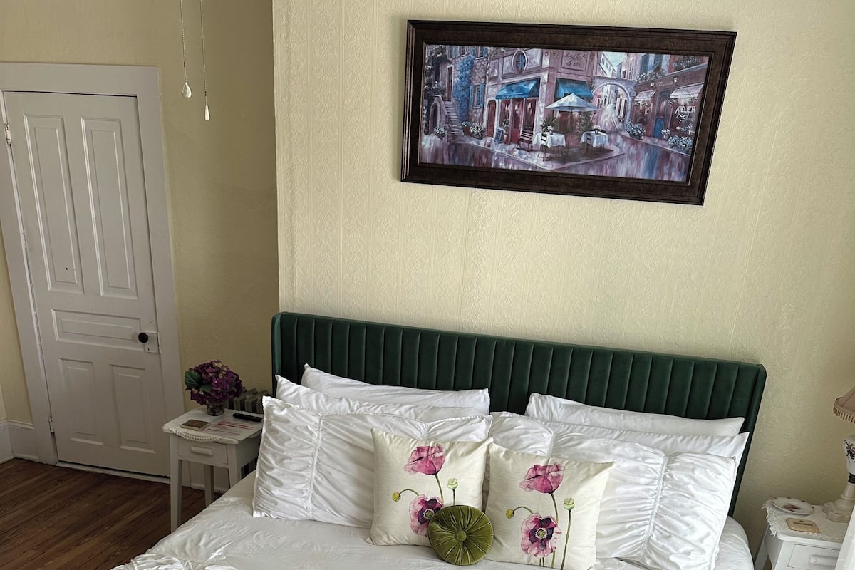 Un lit bien fait, avec du linge de lit blanc et une tête de lit verte, une petite table de chevet ornée d’un vase de fleurs et un tableau encadré accroché au mur.