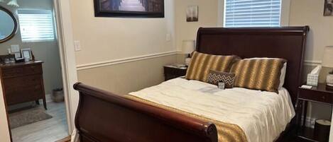 A bedroom with a wooden bed, a nightstand, a dresser, and a window with blinds.