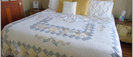 A neatly made bed with a quilt, a wooden headboard, a bedside table with a lamp, and a door in the background.