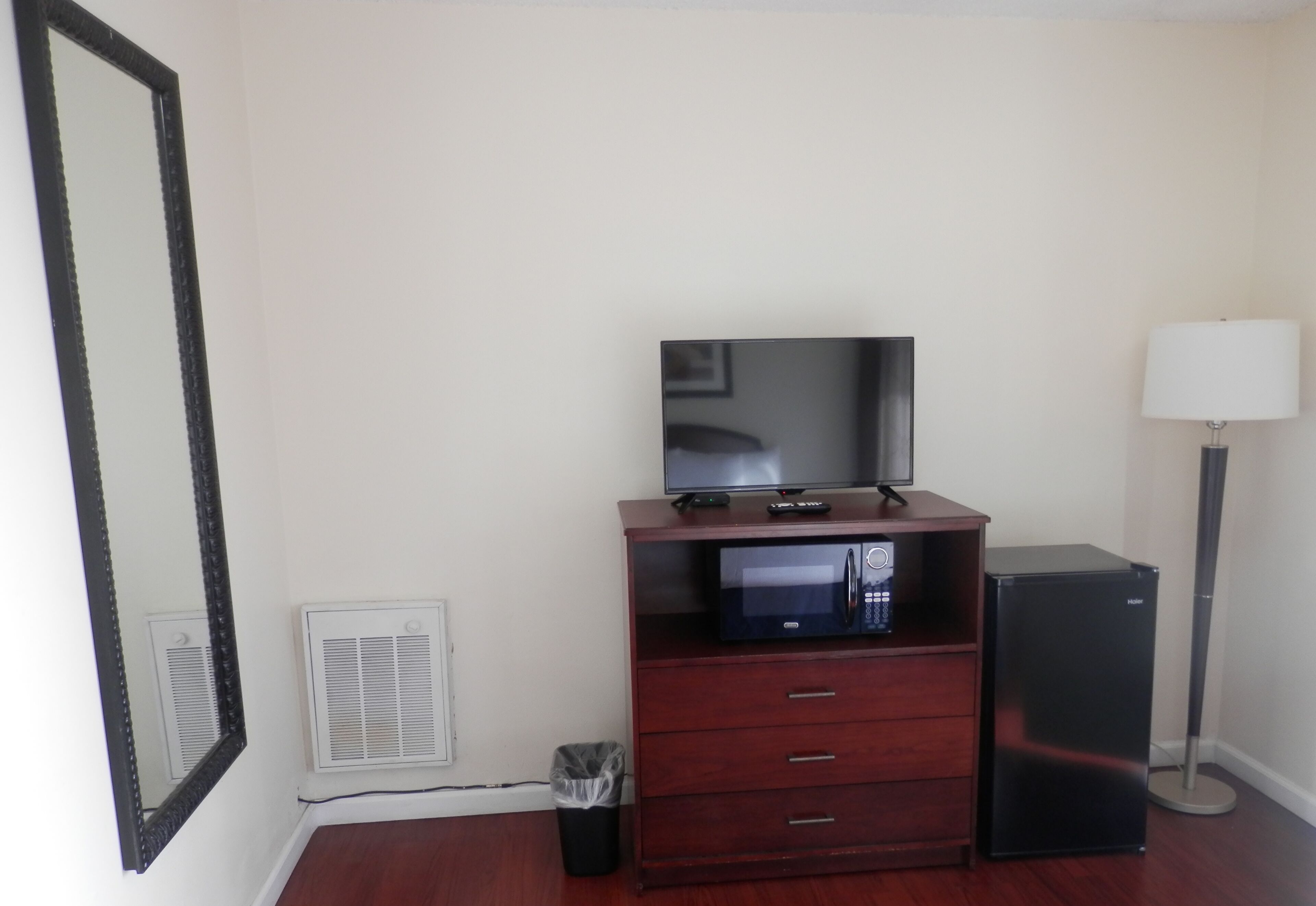 A room with a wooden dresser, a black refrigerator, a microwave, a television, a floor lamp, and a trash can.