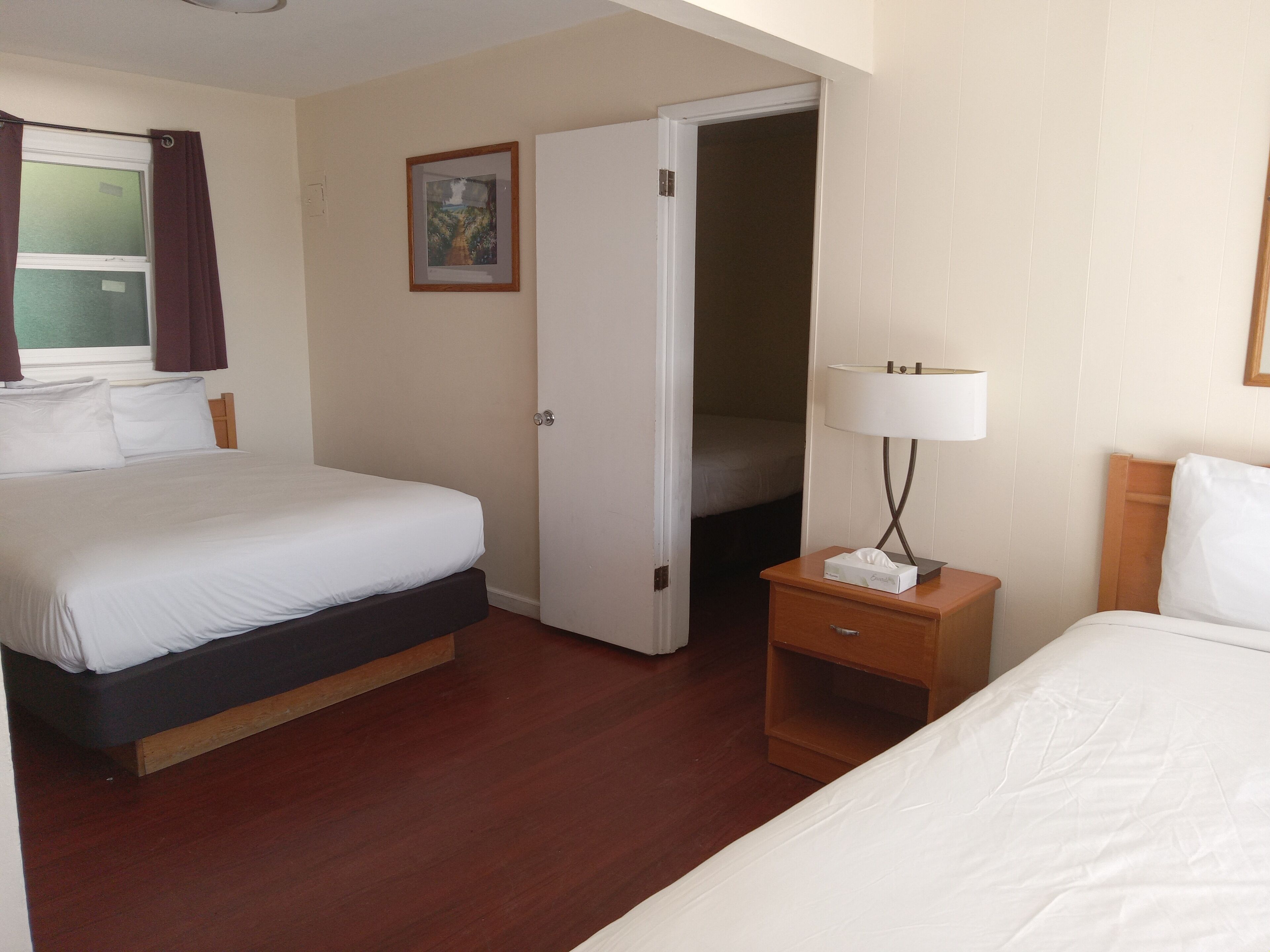 A hotel room with a bed, a bedside table with a lamp, a nightstand, a door leading to another room, and a window with curtains.