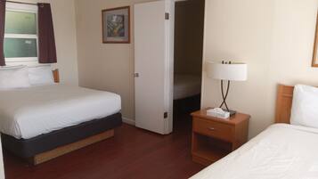 A hotel room with a bed, a bedside table with a lamp, a nightstand, a door leading to another room, and a window with curtains.