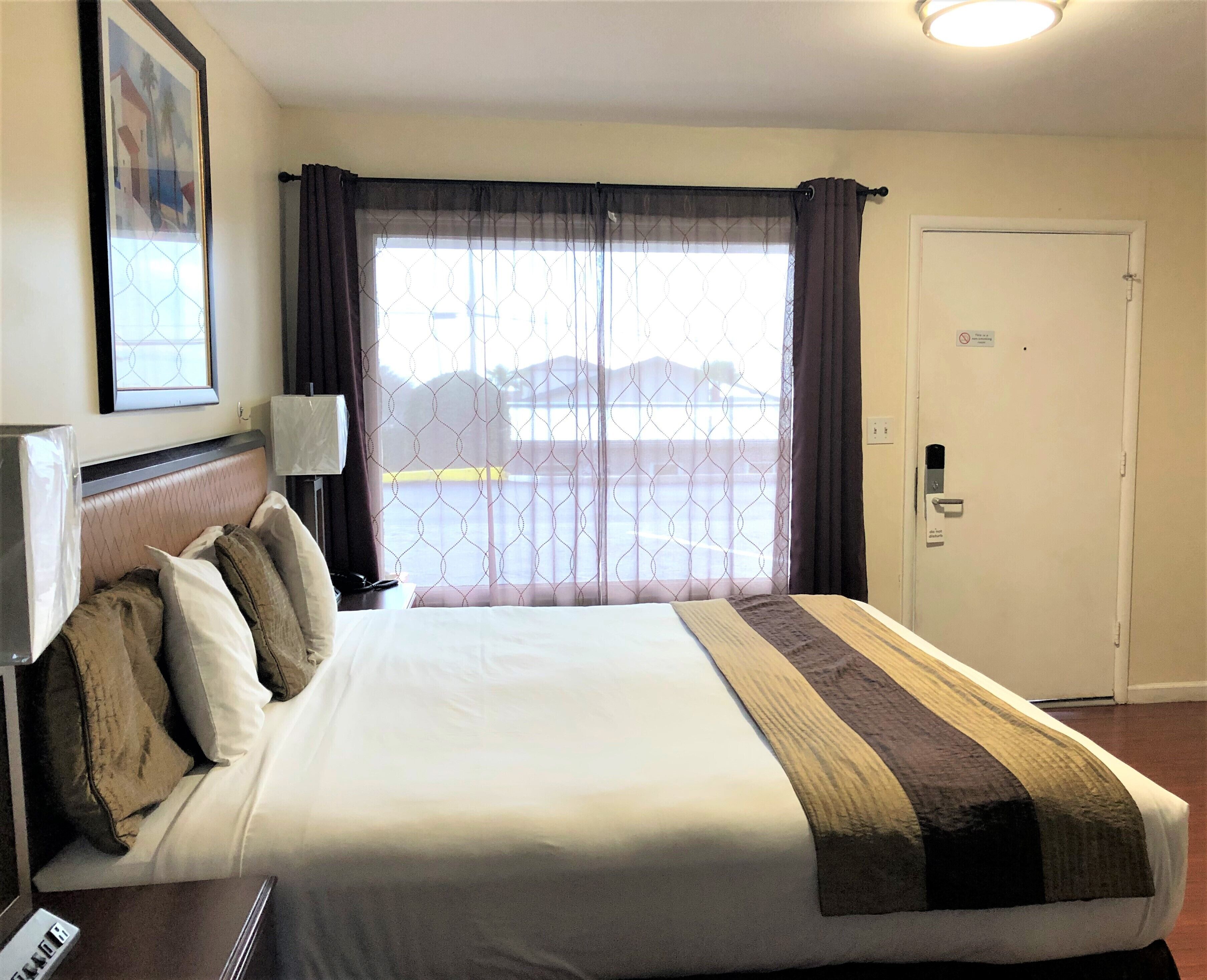 Quarto de hotel com cama, mesa de cabeceira, luminária, porta e janela com cortinas.