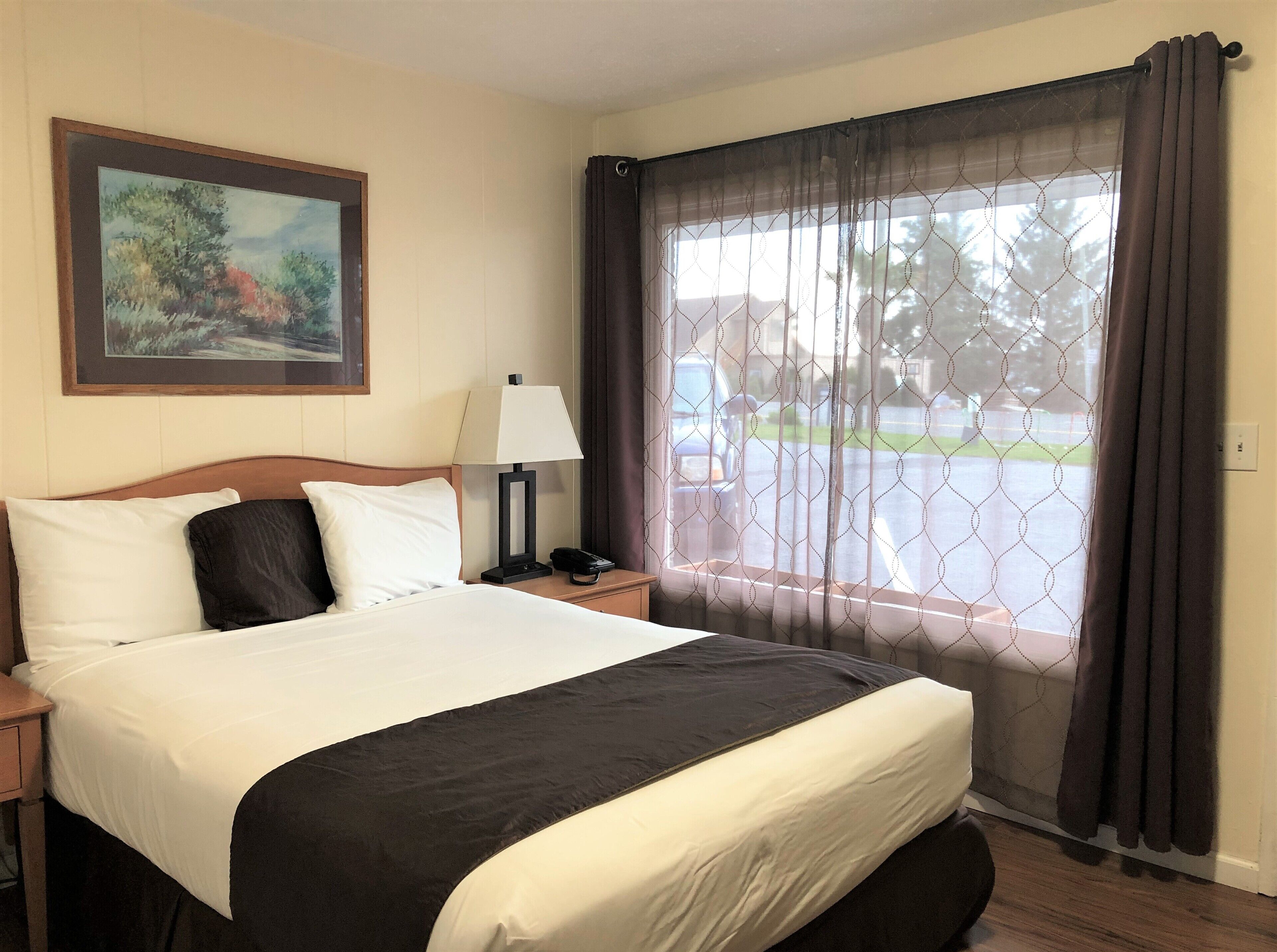 A hotel room with a bed, a nightstand, a lamp, a painting on the wall, and a window with curtains.