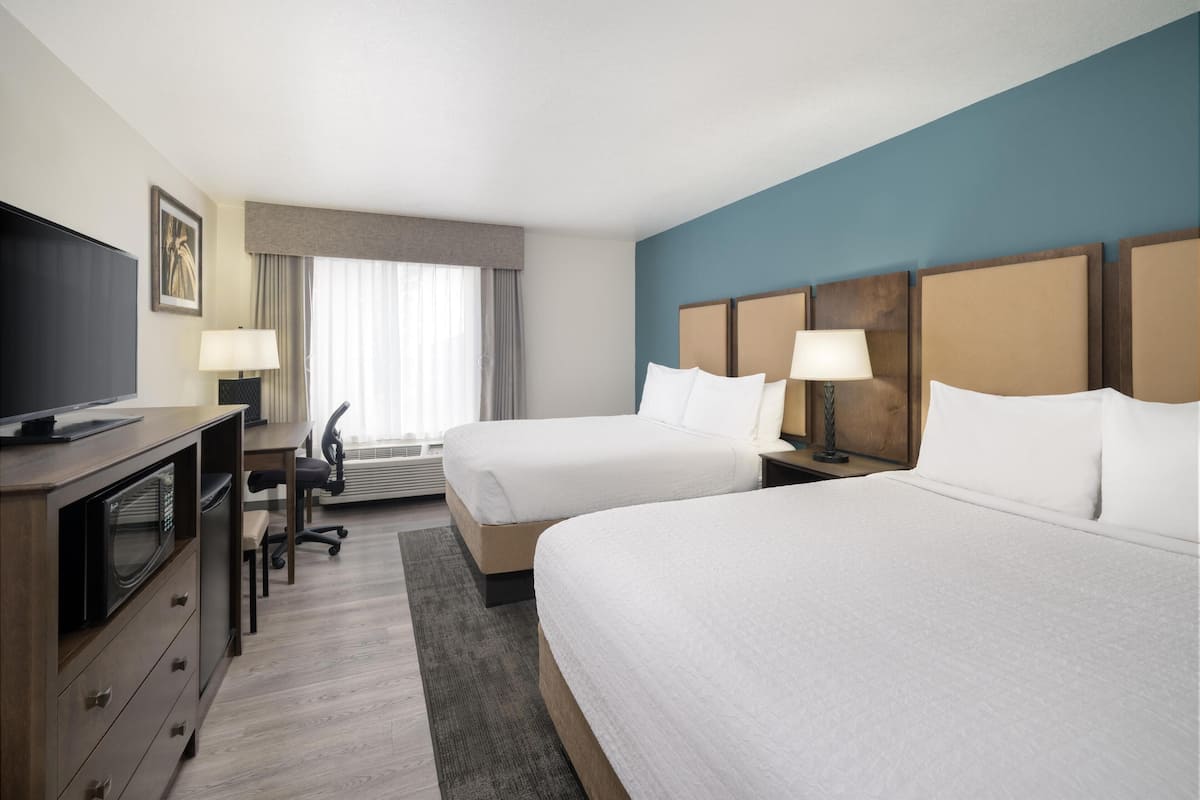 Une chambre d’hôtel avec deux lits, un bureau avec une chaise, un téléviseur à écran plat, une lampe et une grande fenêtre avec des rideaux.