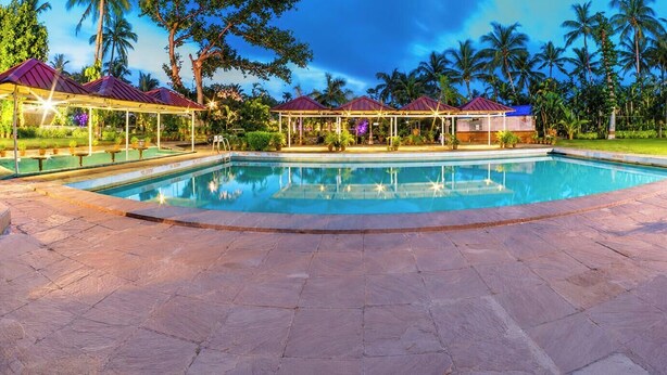 Una piscina con cubierta de piedra, zonas de estar a la sombra y palmeras al fondo.