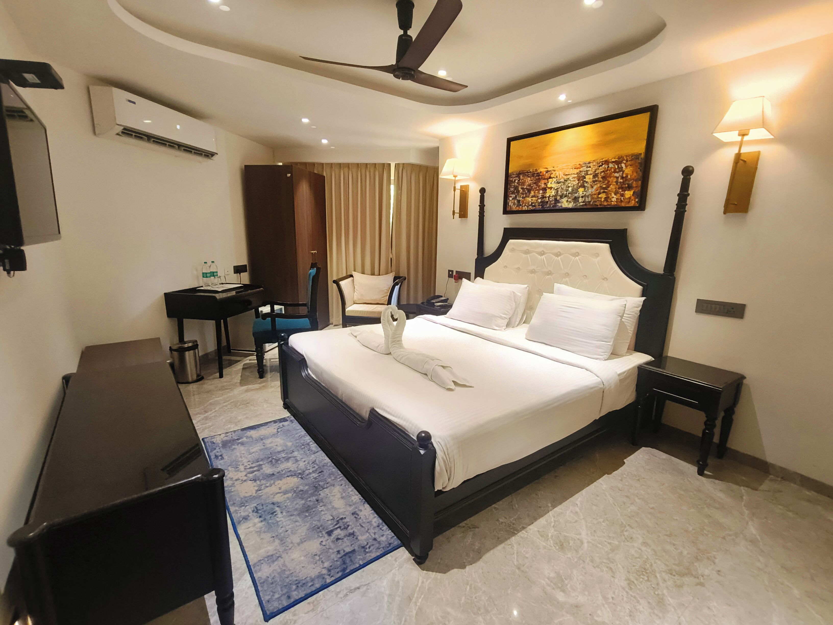 A hotel room with a bed, a desk, a chair, a standing lamp, a ceiling fan, and a painting on the wall.